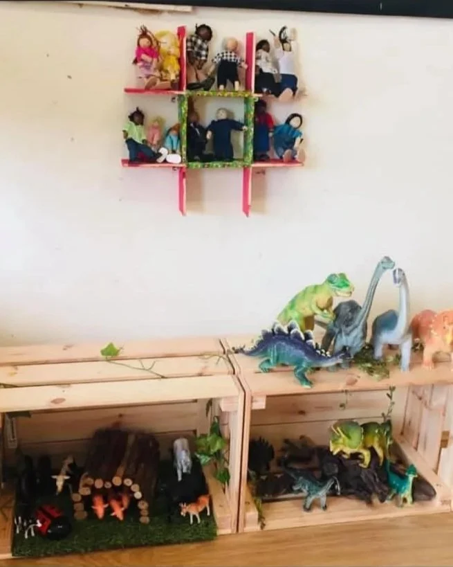 Decorative wooden shelves with plush dolls on the top shelf, various toy dinosaurs on the lower shelves, and some sticks and leaves on the surface.