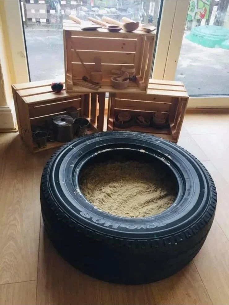 A large car tire filled with sand, placed on a wooden floor next to wooden crates filled with small bowls and cups, near a glass door.