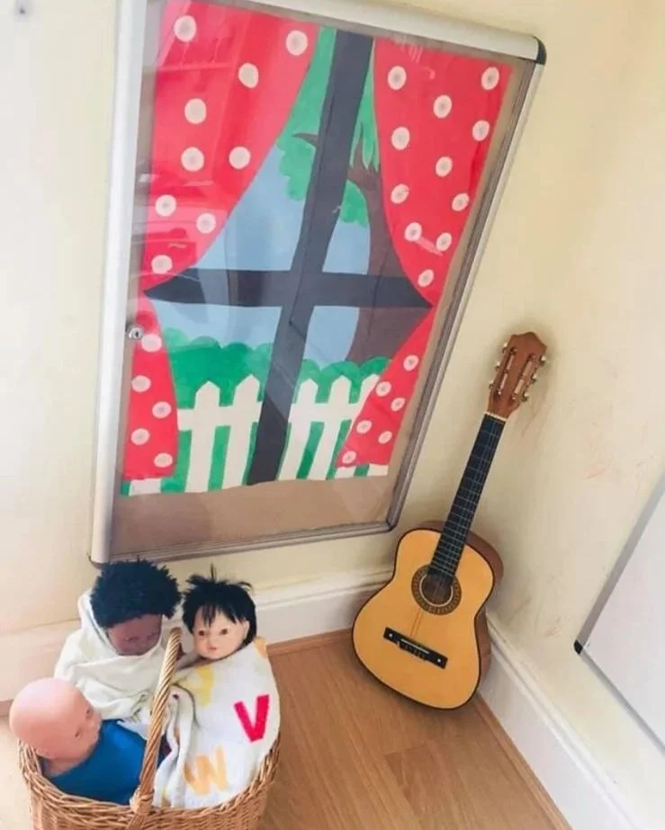 A children's play area with a small basket containing three dolls and a blanket, a wooden guitar leaning against the wall, and a window with colorful curtains that have a tree and fence design.