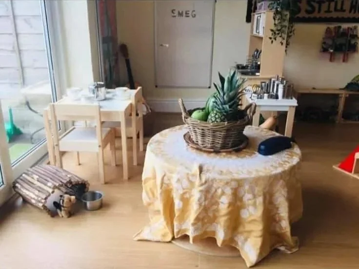 A cozy indoor space with a round table covered with a yellow tablecloth holding a basket of fruit, including a pineapple, an apple, and a banana. To the left, there are two small chairs, a dog bed, and a dog near a metal bowl. In the background, ther