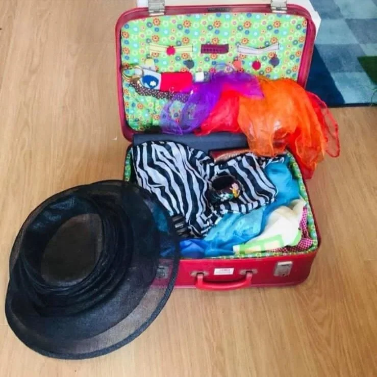Open red suitcase filled with colorful clothes and accessories, with a black wide-brimmed hat placed in front of it on a wooden floor.
