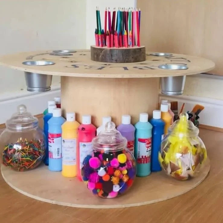 A wooden circular craft caddy with two tiers, holding various art supplies. The top tier has a round wooden holder with colorful pens, while the bottom tier contains bottles of paint, jars filled with push pins, pom-poms, and folded paper, along with