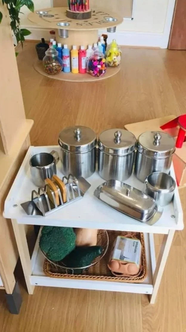 A white trolley with metal containers, measuring cups, and utensils on top, and a basket with a green loofah, cucumber, and soap at the bottom, in a room with wood flooring and shelves with colorful bottles and decorations.