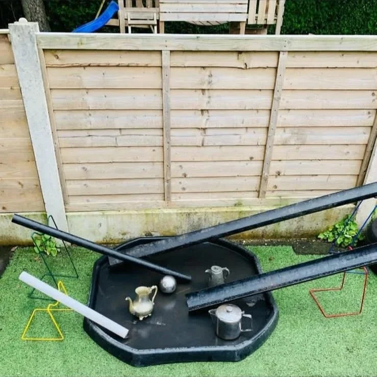 A small backyard with a wooden fence, artificial grass, and a black plastic kiddie pool placed on the grass. The pool is filled with water and contains various objects like a teapot, a metal ball, and some black pipes or rails resting across it. Ther