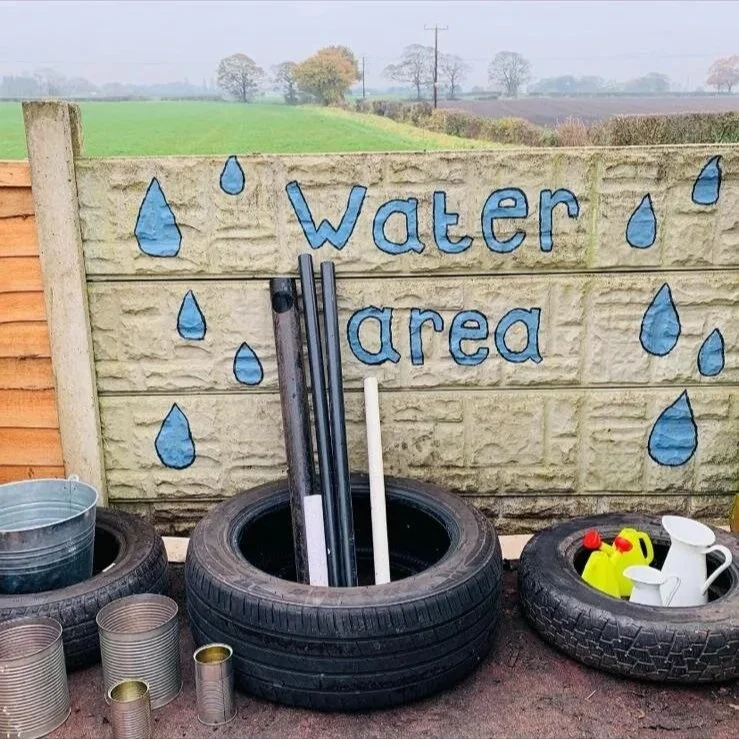 Sign reading 'Water area' with blue water droplets on a wooden fence, surrounded by tires filled with gardening tools and supplies like watering cans and buckets, outdoors on a sunny day with fields and trees in the background.
