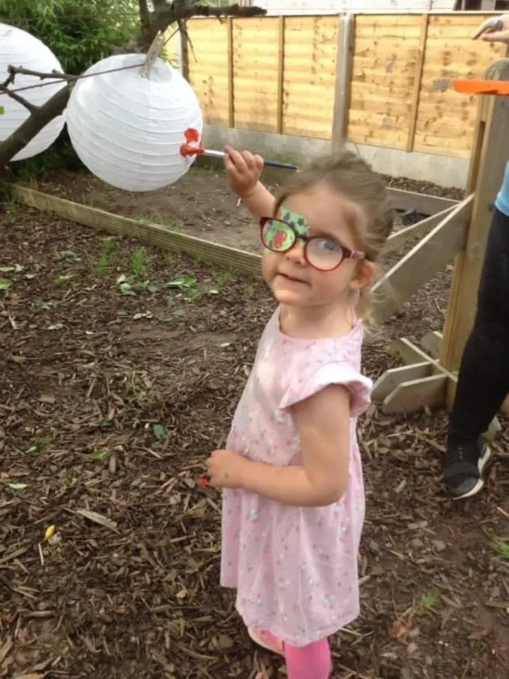 Child holding a paintbrush next to a white paper lantern hanging from a tree in a backyard.