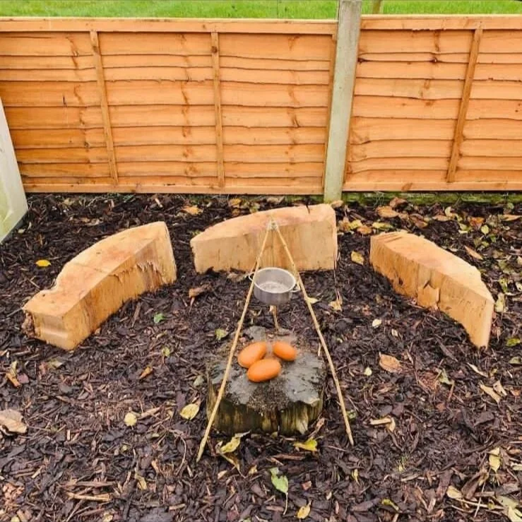 A garden with wooden bench-like structures in a semi-circle around a tree stump, which has four eggs and a small pot hanging above it, supported by thin wooden sticks, on a bed of dark mulch with a wooden fence in the background.