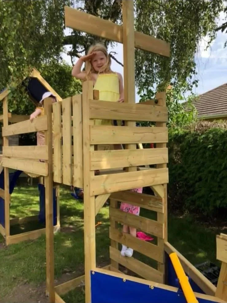 Two young girls playing on a homemade wooden play structure in a backyard, surrounded by trees and bushes.