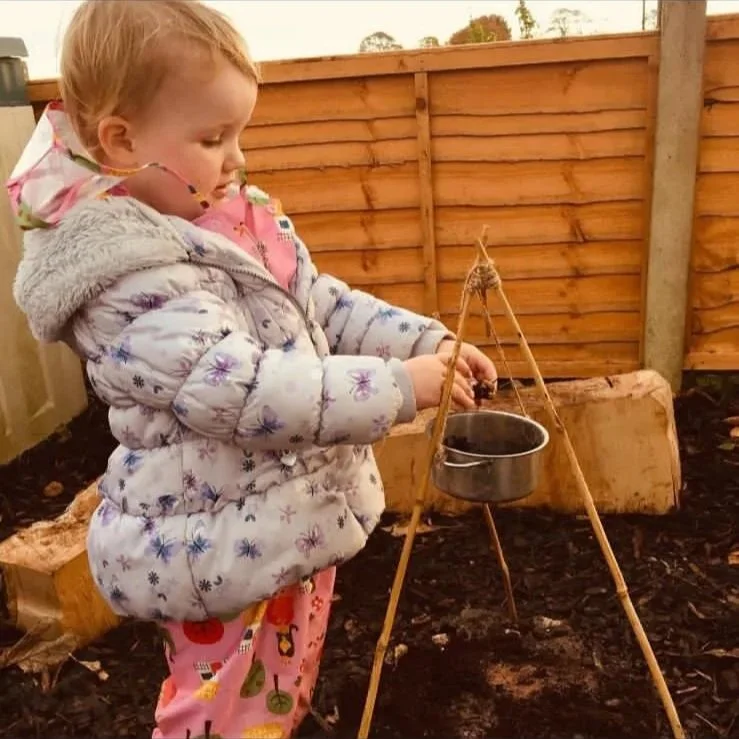 A young girl with blonde hair, dressed in a winter jacket and pink pajamas, is standing outdoors near a wooden fence. She is engaged in an activity involving a small pot suspended over soil, supported by a tripod made of sticks, suggesting gardening 