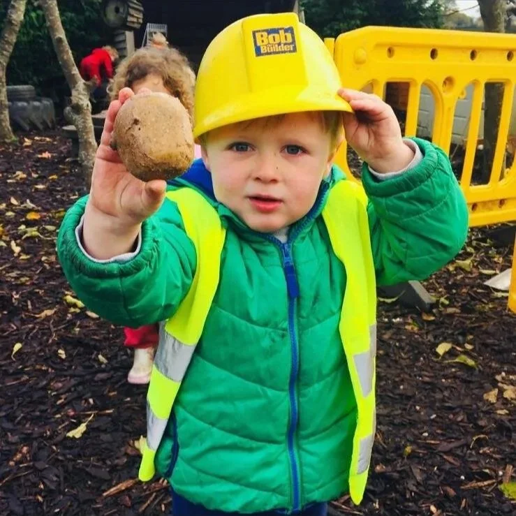 A young boy wearing a yellow construction helmet labeled 'Bob the Builder', a neon yellow safety vest, and a green jacket, holding a large potato in his right hand and looking at the camera.