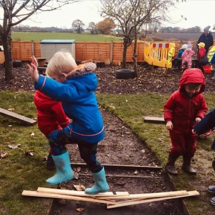 Kids playing outdoors on a muddy path with wooden planks, one child in blue boots balancing on the planks in front of others in a garden or playground area, with trees and other children in the background.