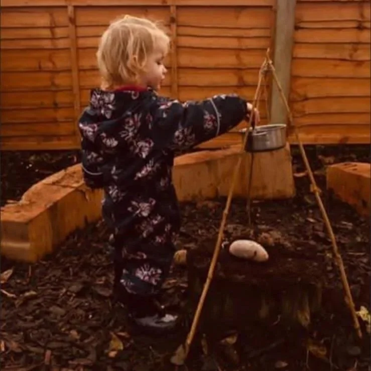 Child with blonde hair in a patterned navy jacket and pants, standing outdoors next to a wooden fence, playing with a hanging kettle and a rock beneath it, outdoor setting with wood chips on the ground.