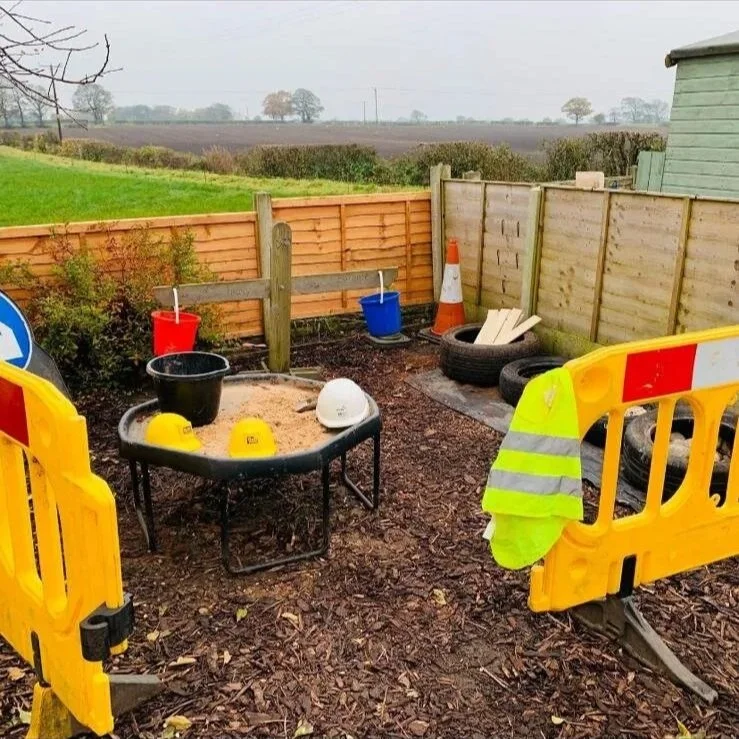 Construction site in a backyard with yellow safety barriers, a children's sandbox with yellow, white, and black helmets, buckets, a white hard hat, a traffic cone, in-ground tires, and a wooden fence with a view of fields in the background.
