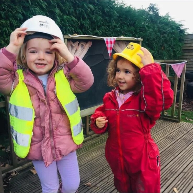 Two young girls dressed in construction gear, wearing helmets and high-visibility vests, standing outdoors on a wooden surface with festive bunting in the background.