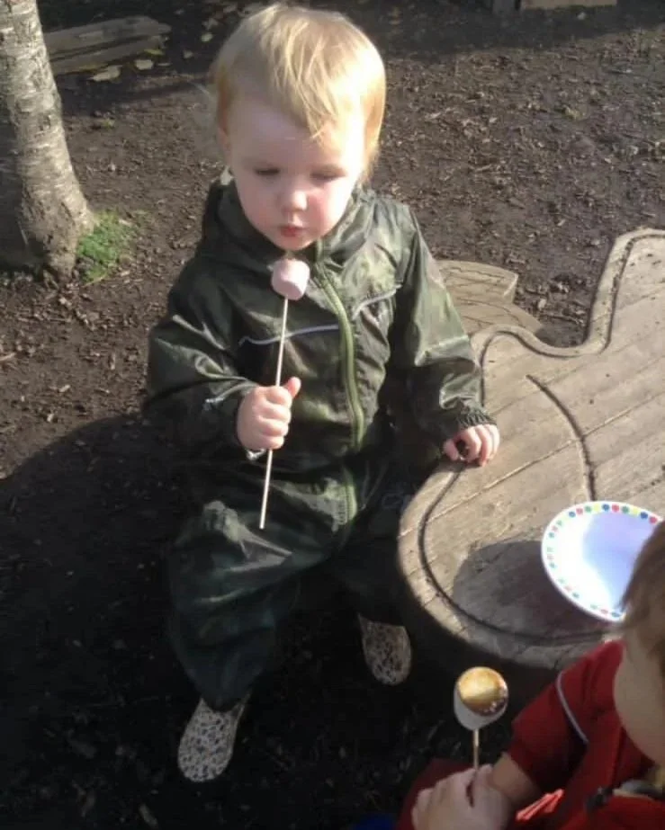 A young child with blonde hair wearing a green jacket and camouflage boots sits on a wooden park bench holding a pink lollipop. Another child in red is partially visible, also holding a lollipop. The ground is dirt, and there is a tree nearby.