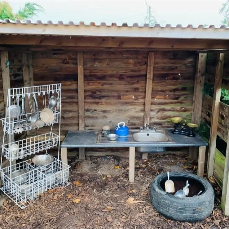 Outdoor wooden kitchen setup with a small sink, a blue kettle, a stove with two yellow burners, a white dish rack with kitchenware, and a tire used as a container holding gardening tools, all under a corrugated metal roof.