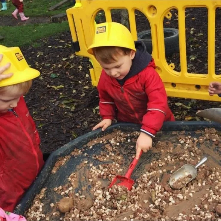 Two young children wearing yellow hard hats and red jackets playing in a sandbox with a small shovel and a toy trowel, surrounded by a yellow plastic fence outdoors.