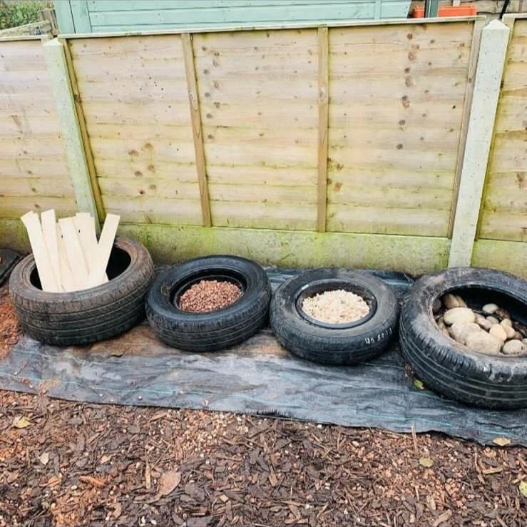 Four recycled tires used as planters against a wooden fence, containing wooden sticks and various rocks, on a tarp with wood mulch on the ground.