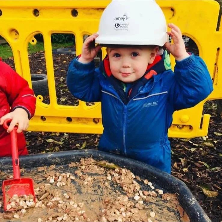 Child wearing a white safety helmet and blue jacket, standing outdoors with playground equipment and leaves on the ground.