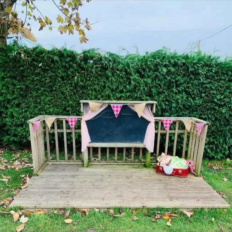 Empty outdoor wooden deck with a small railing and a hedge in the background. There is a decorated chalkboard or signboard with pink curtains and bunting flags hanging on it. To the right, on the deck, there is a small red suitcase filled with bags a