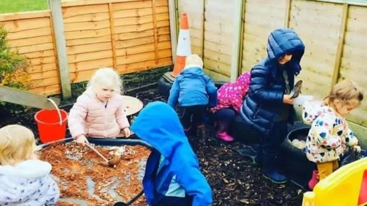 Children playing in a sandbox and on a tire swing in a fenced outdoor play area.