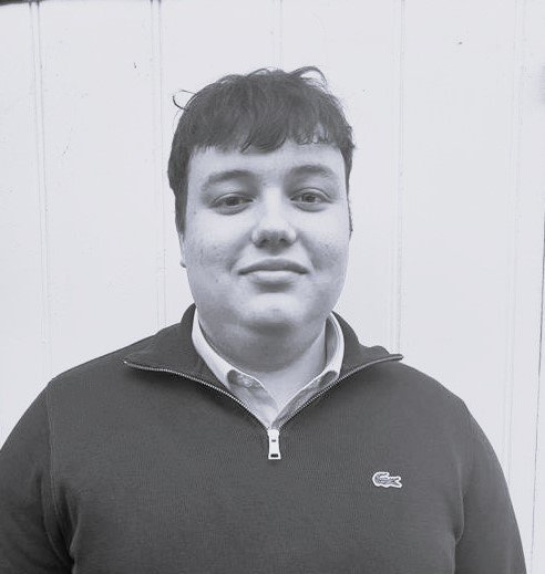 A young man with short dark hair wearing a dark zip-up jacket over a collared shirt, standing indoors against a plain wall.