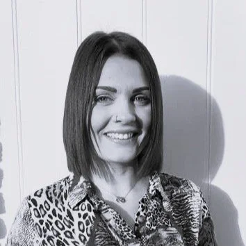 Black and white photo of a woman with shoulder-length dark hair and a nose piercing, smiling and wearing a patterned top, standing in front of a wooden wall.