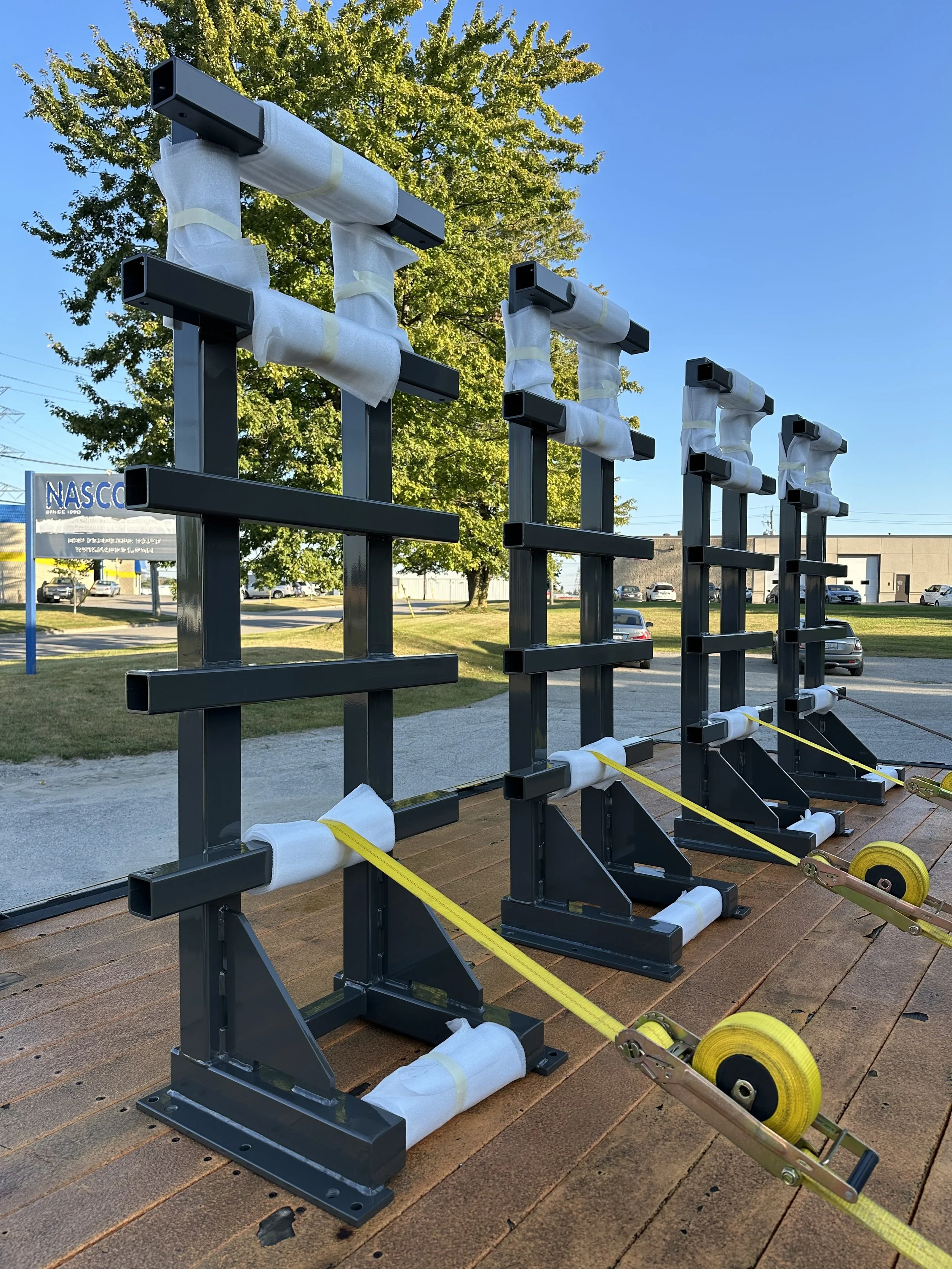 powder coated steel frames, Strapped on a trailer for delivery, with trees, cars, and a NASCO sign in the background, under a clear blue sky.