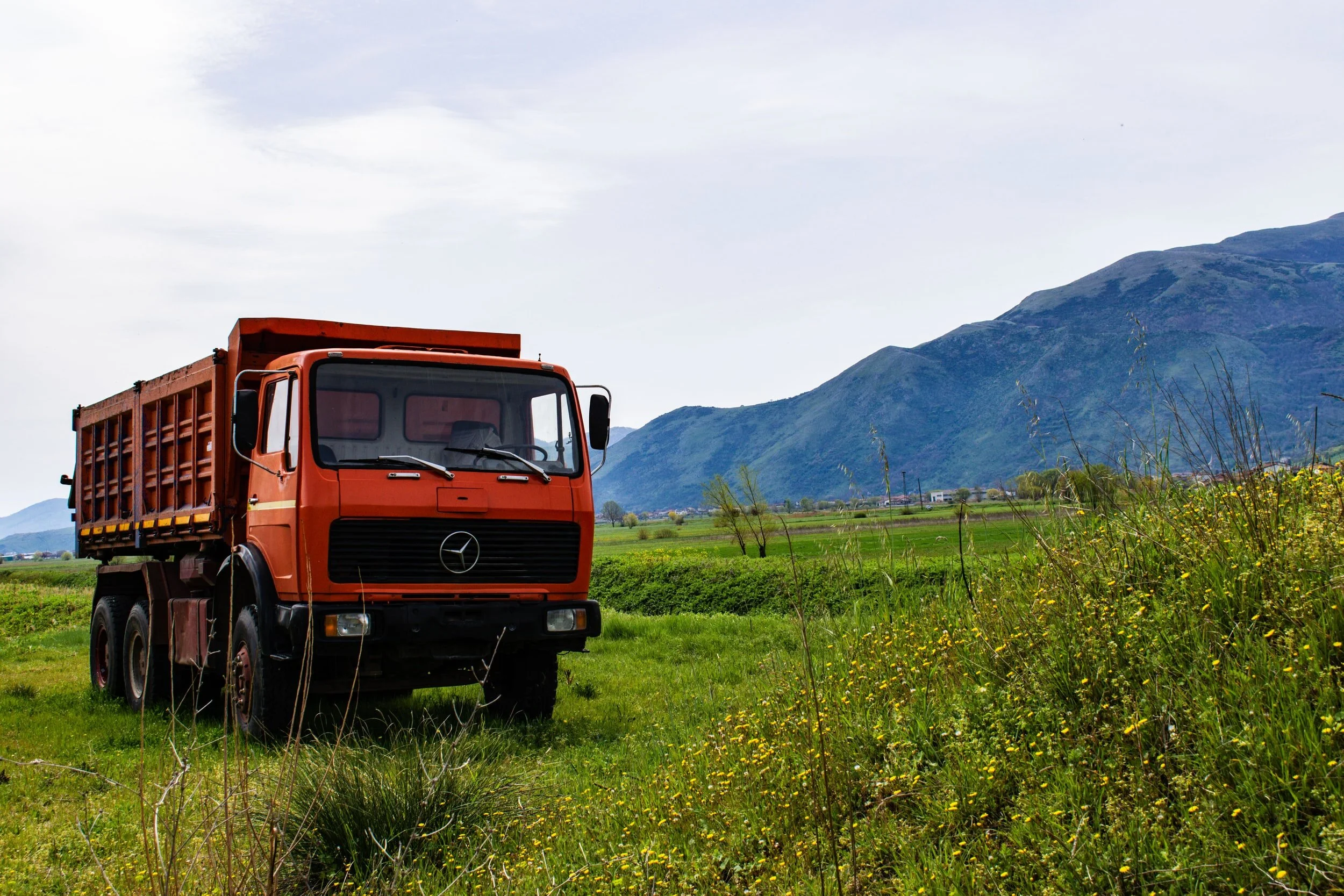 Orange Mercedes-Benz dump truck parked on grassy field with mountains in the background under a cloudy sky.