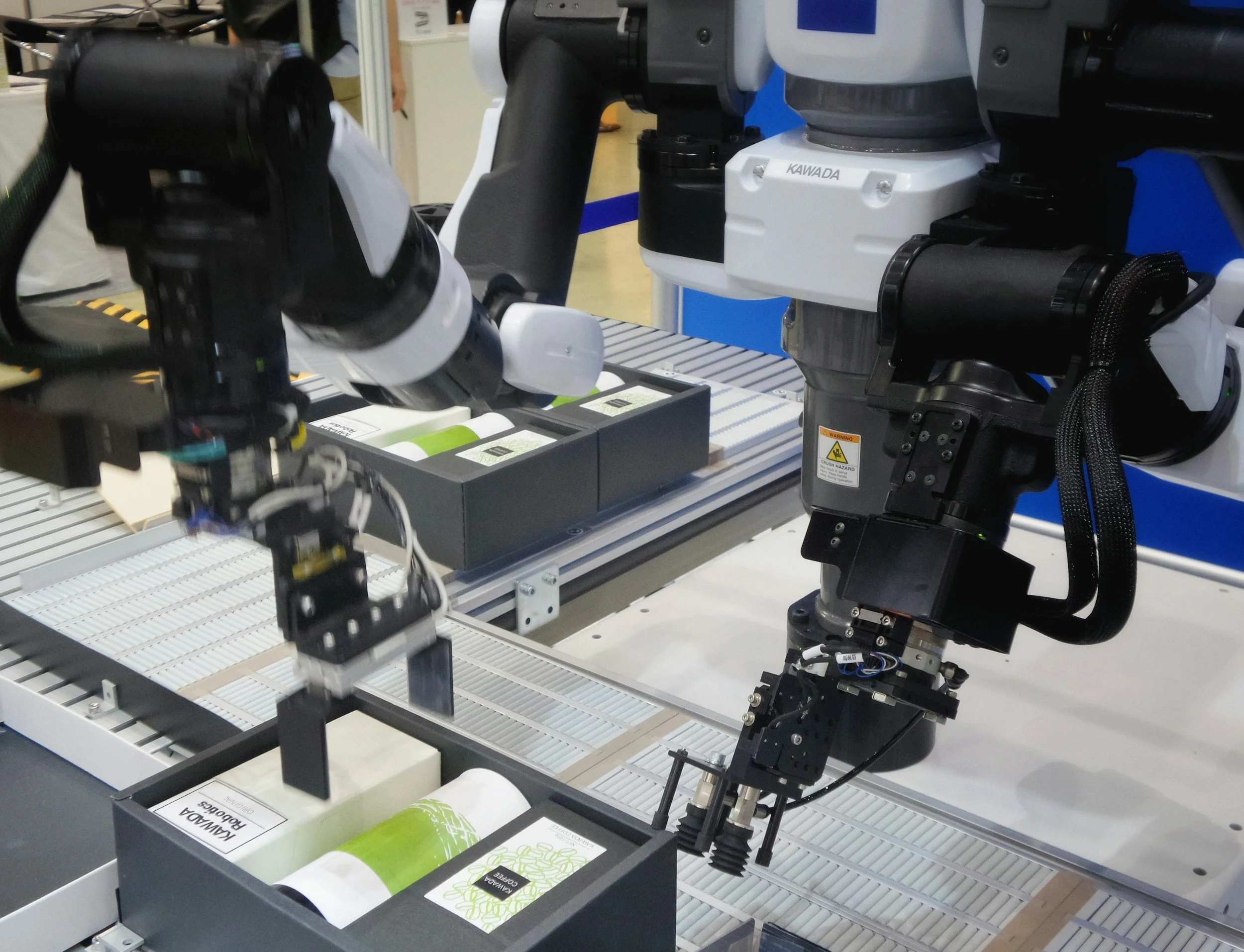 Two robotic arms working on packaging or inspecting cylindrical products on a conveyor belt in a manufacturing or industrial setting.
