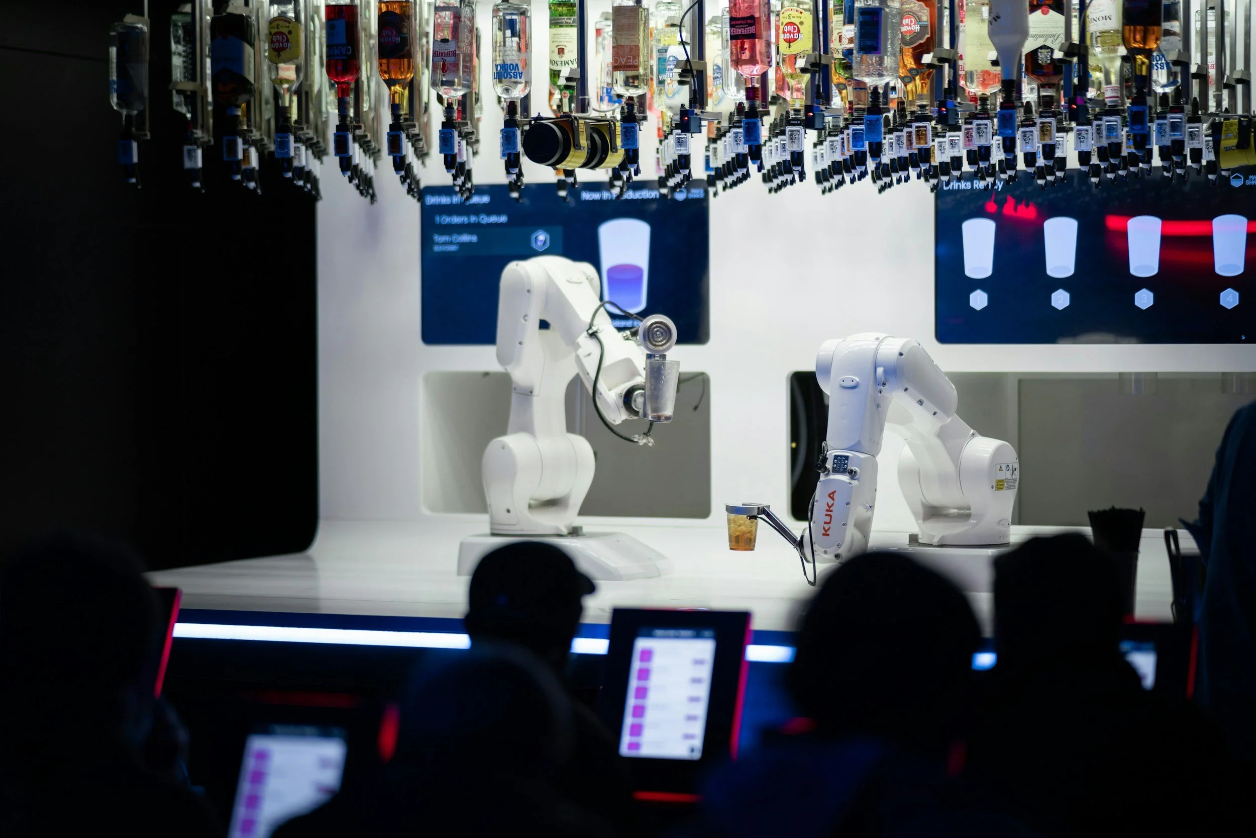 Two white industrial robots on a lab bench with red and orange containers attached to them. There are people in dark clothing with illuminated screens in the foreground, and digital screens in the background showing a purple glass and an exclamation mark alert.