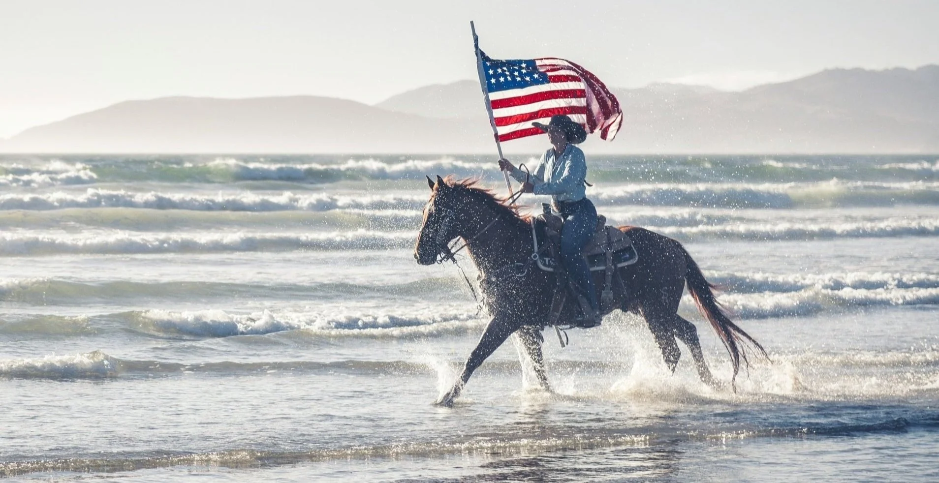 Person riding a horse in the shallow ocean water while holding an American flag, with mountains in the background.