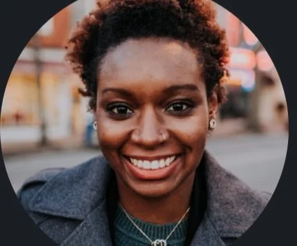 Close-up of a smiling woman with short curly hair, wearing a dark jacket and a necklace, standing outdoors in an urban setting.