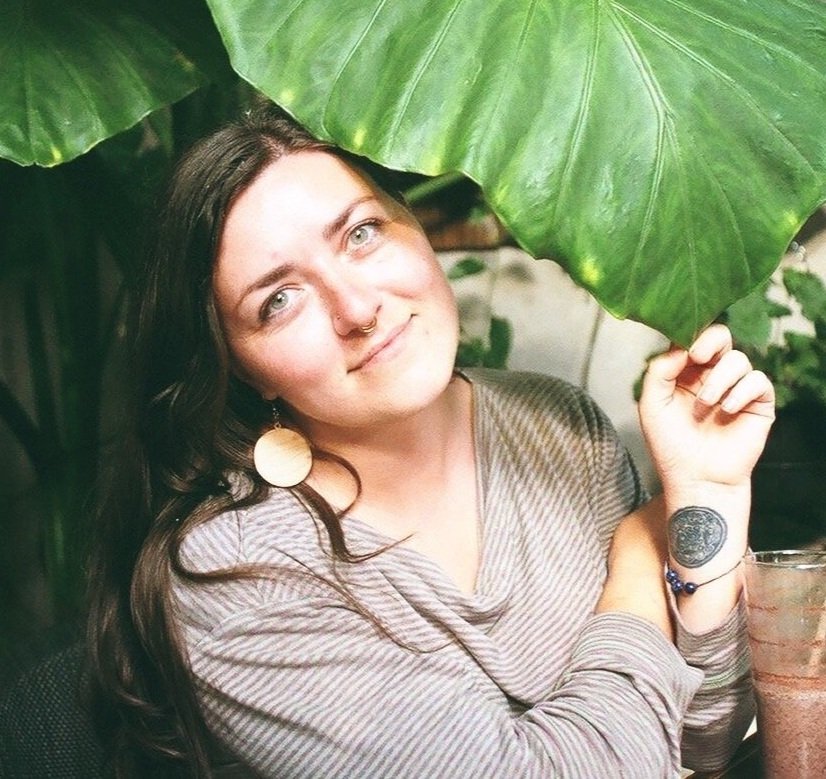 A young woman with long dark hair, wearing large round earrings, a striped shirt, sitting behind large green leaves, holding one leaf above her head, smiling softly, with a drink nearby.