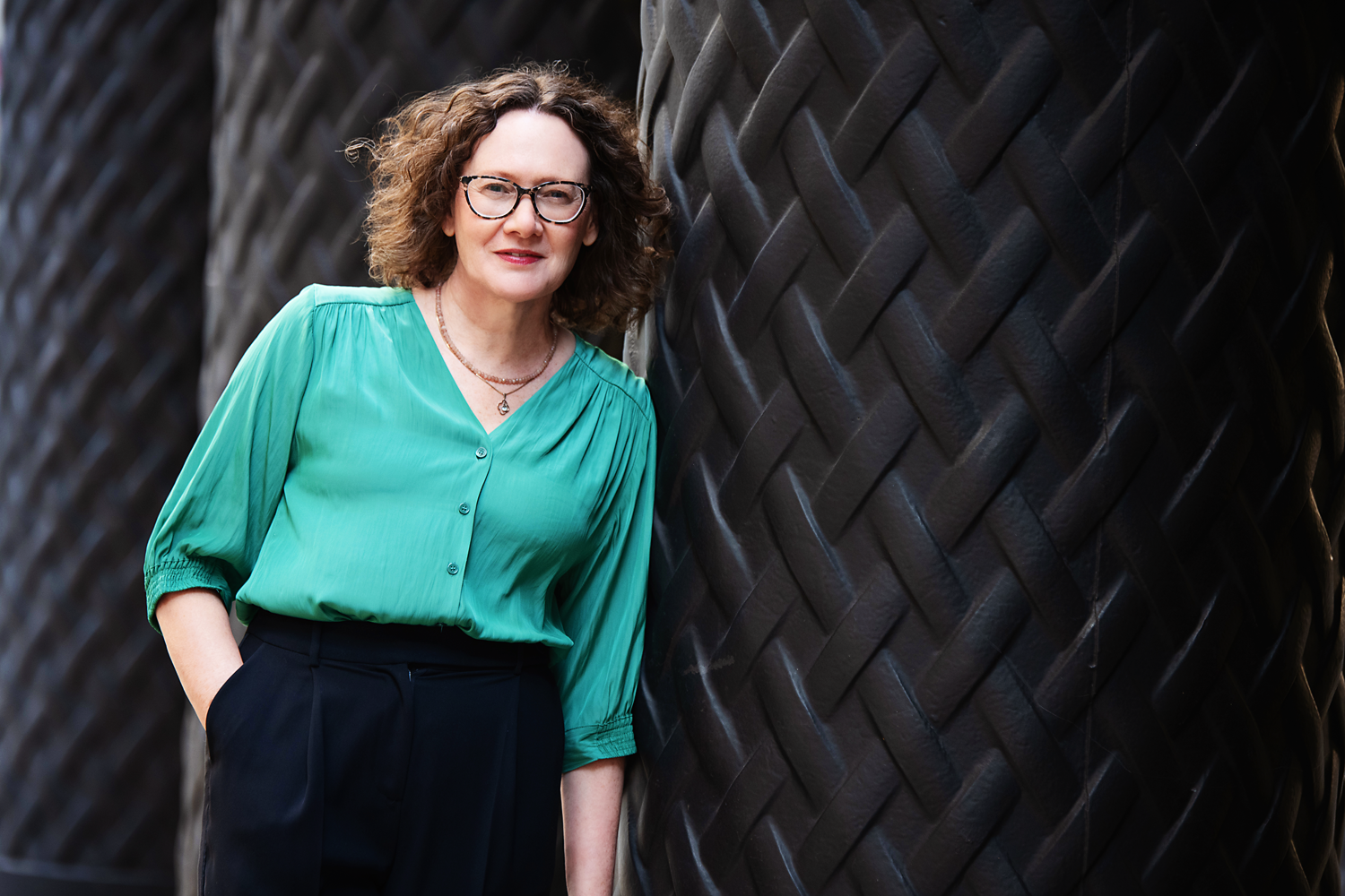 Emma Marinos wearing a green blouse and black pants standing next to a large black sculpture with a textured, patterned surface.