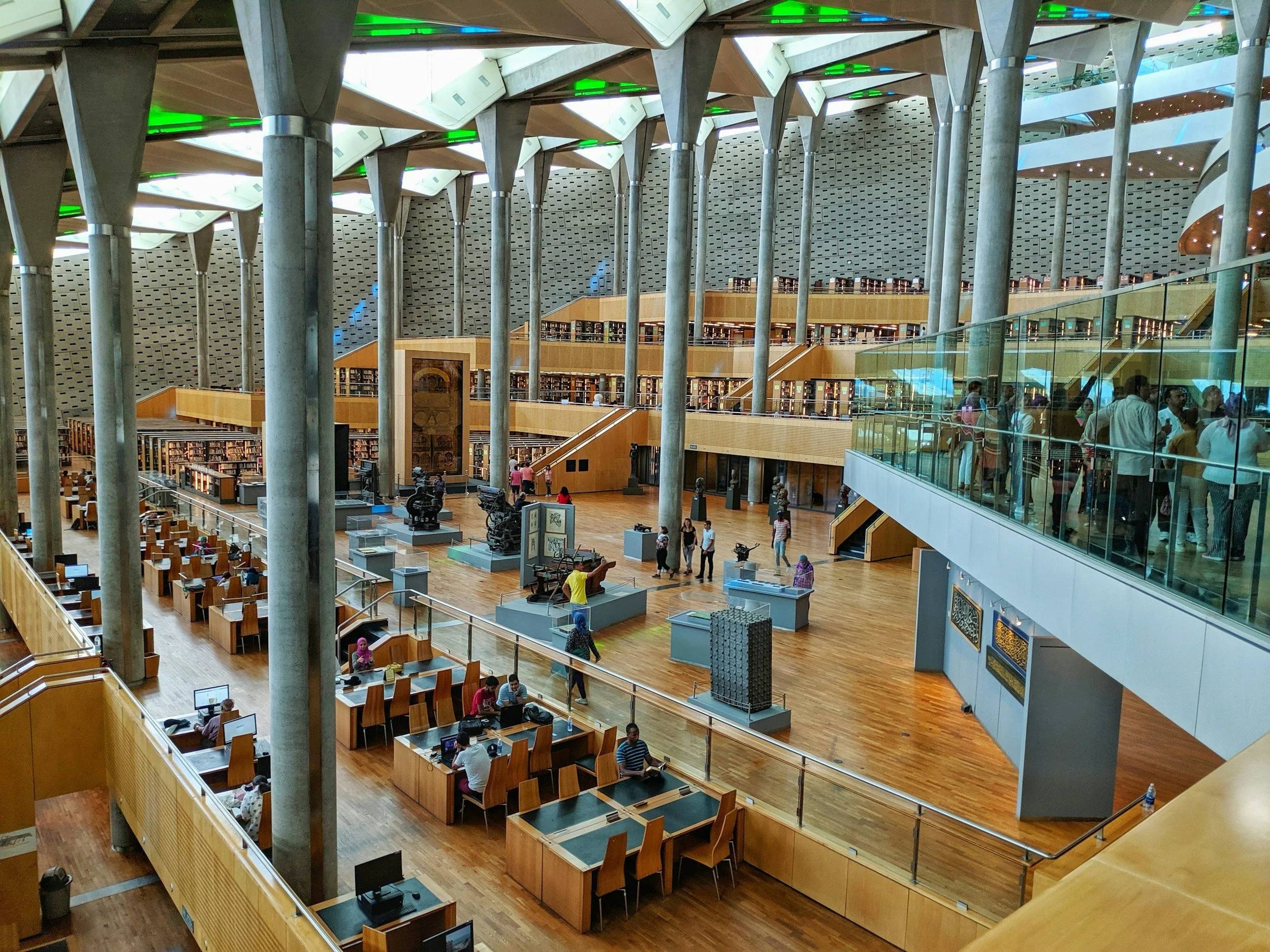 Interior view of a modern multi-level library with wooden desks, bookshelves, and people studying and walking around. Steel columns support the ceiling with large windows and green accents.