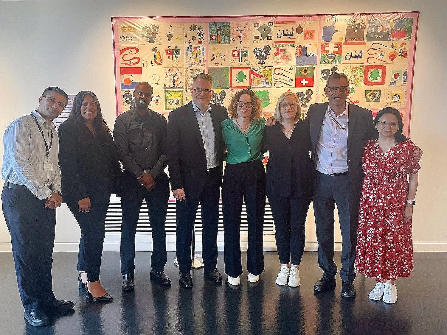 Group of nine diverse people standing together in front of a colorful artwork on a wall. They are dressed in business casual and formal attire, smiling at the camera.