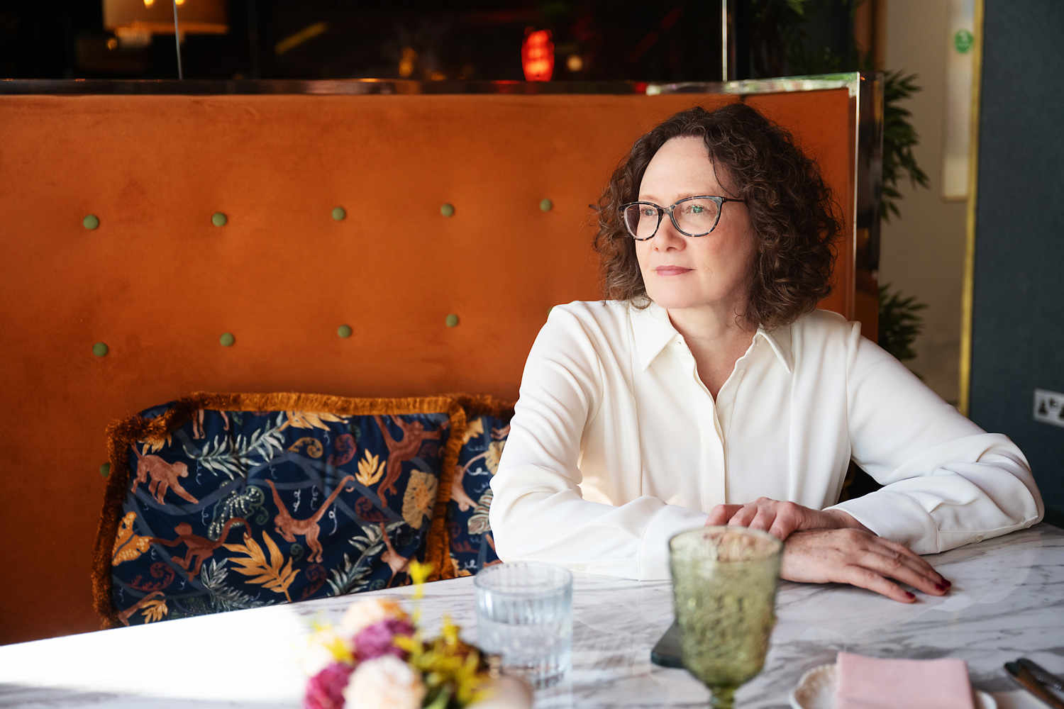 Emma Marinos sits at a marble table in a restaurant, wearing a cream-colored blouse, looking thoughtfully to the side.
