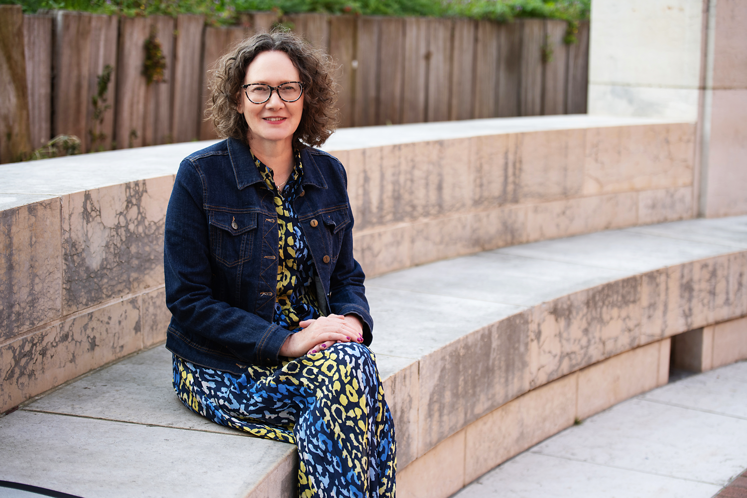 Emma Marinos sitting on a curved stone bench outdoors, smiling at the camera, wearing a denim jacket and a colorful patterned dress.