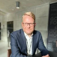 A man with glasses in a suit sitting at a table in an office or conference room.