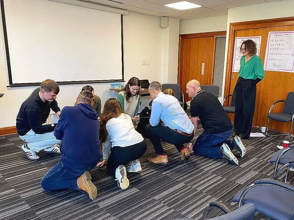 Group of people kneeling on the carpeted floor and looking at something in the middle, in what appears to be a conference or meeting room.