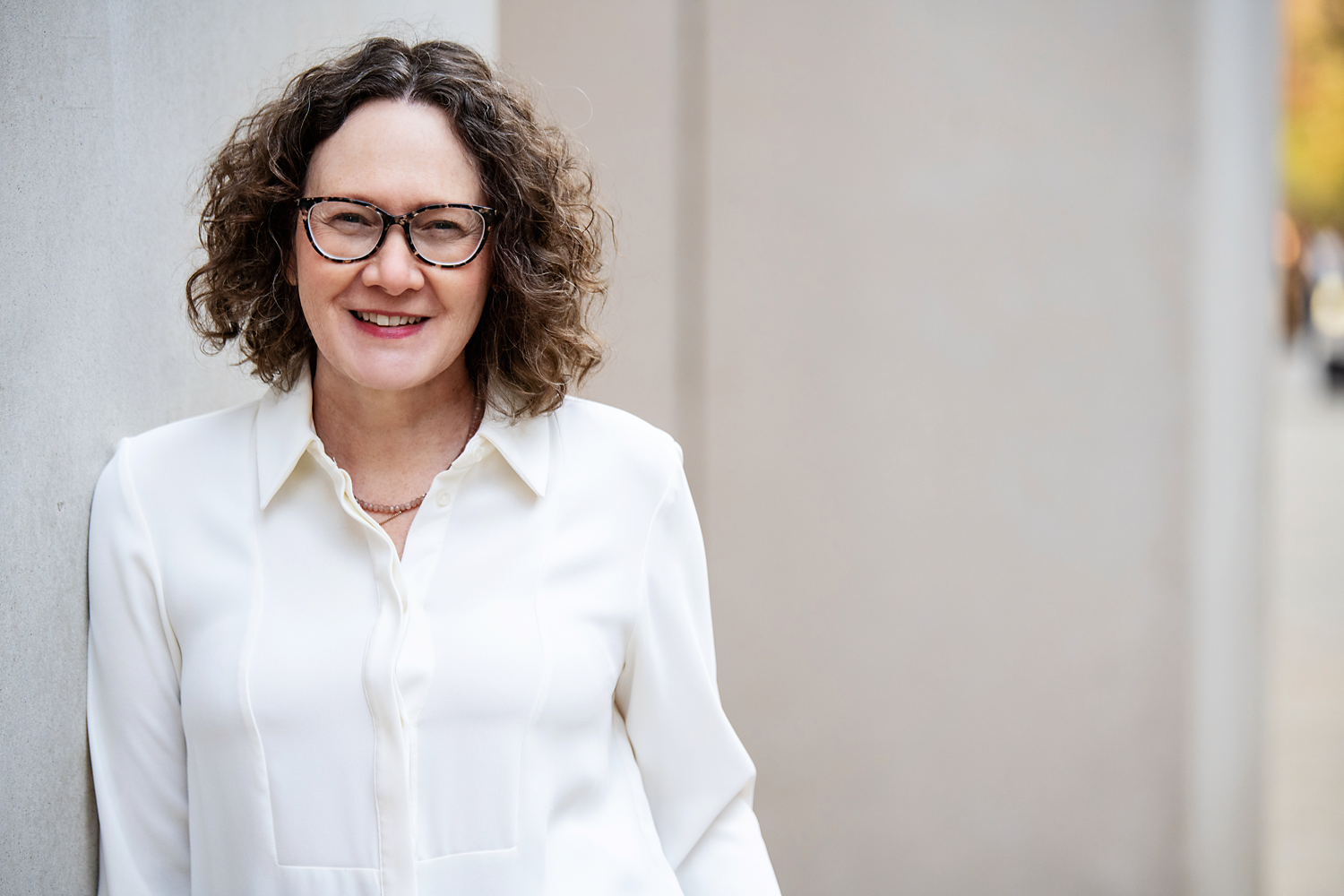 Emma Marinos in a white blouse smiling while leaning against a concrete wall outdoors.