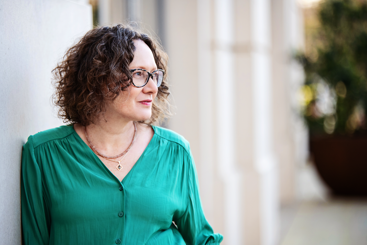 Emma Marinos in a green blouse standing near a wall with a soft focus background.