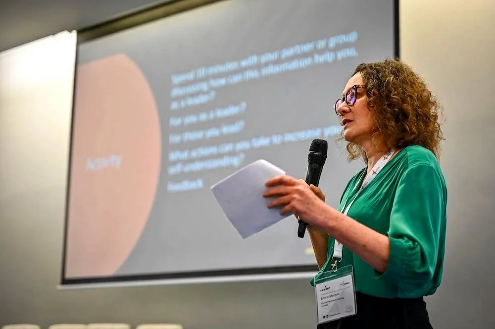 Woman in green blouse holding a microphone and notes, speaking at a conference with a presentation slide visible in the background.