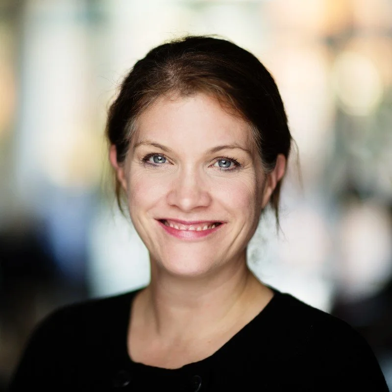 Close-up portrait of a woman with a warm smile, dark hair, and blue eyes, wearing a black top, with a blurred, colorful background.