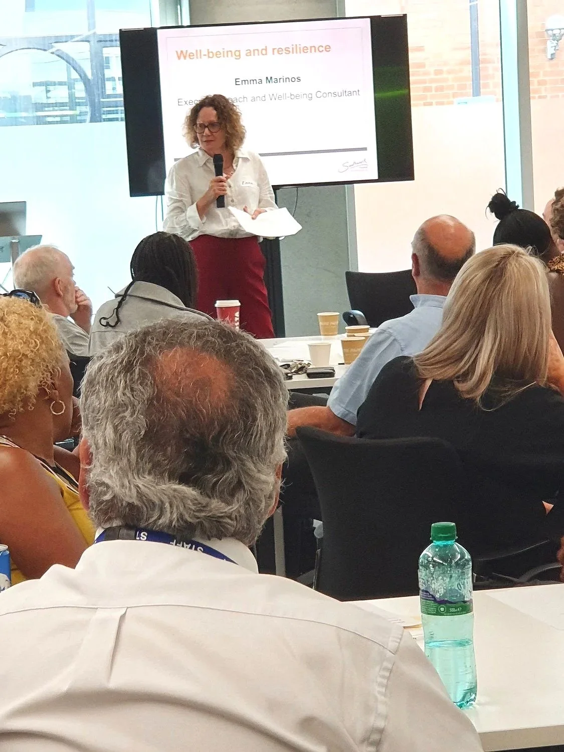 A woman giving a presentation on a topic called 'Well-being and resilience' to an audience in a conference room. The presentation slide displays her name, Emma Marinos, and her title as an Executive Coach and Well-being Consultant. She is holding a microphone and standing in front of a large screen.