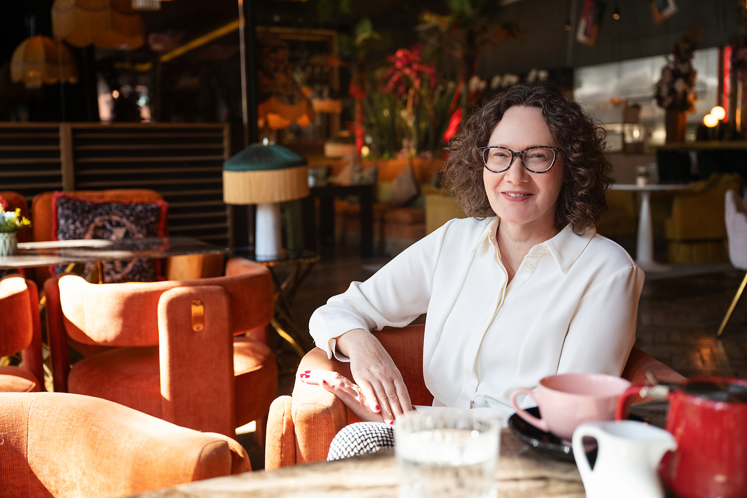 Emma Marinos in a white shirt sitting at a table in a cozy, well-lit cafe. There are teapots, cups, and a glass of water on the table. The background includes warm-colored chairs, lamps, and decorative plants.