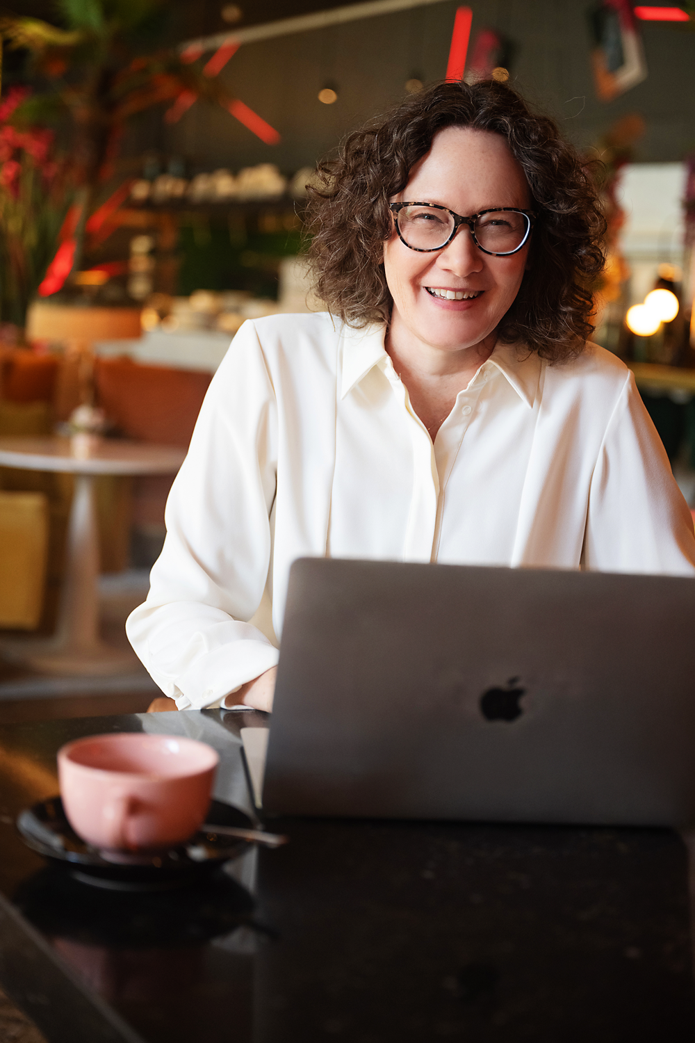 Emma Marinos smiling at a laptop in a cozy, warmly lit cafe.