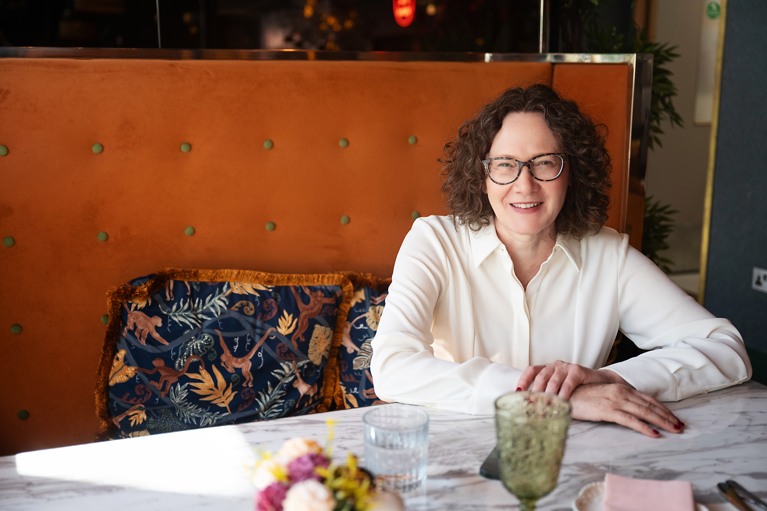 Emma Marinos sitting at a marble table, smiling, in a restaurant or cafe setting.