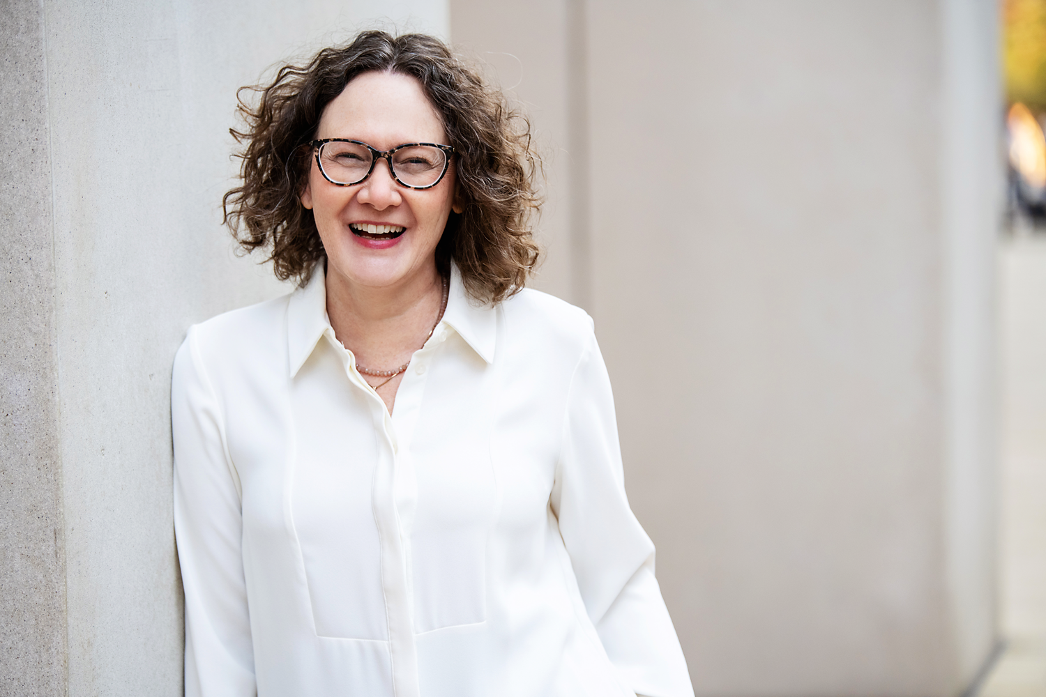 Emma Marinos in a white blouse, smiling and leaning against a concrete wall outdoors.